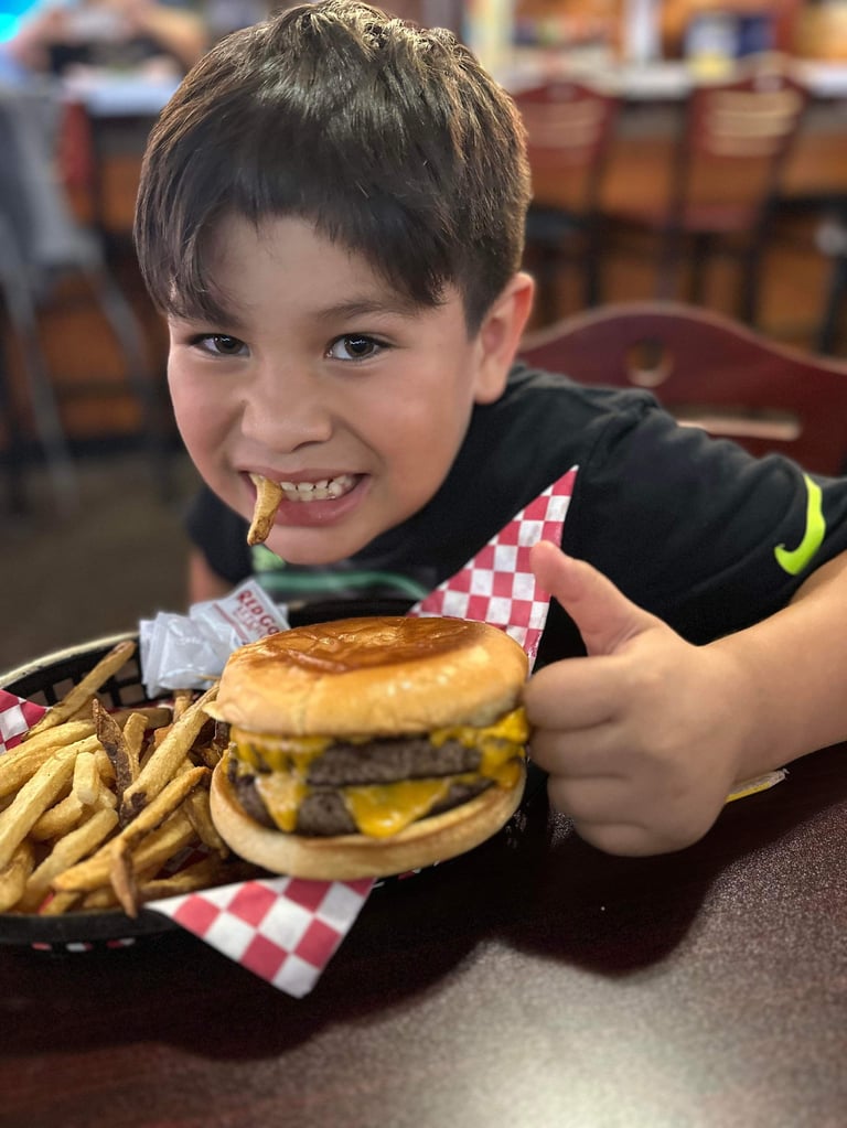 Young boy smiling while holding a cheeseburger and fries on a checkered paper at a restaurant