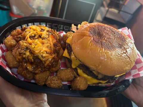 A cheeseburger with melted cheddar and crispy tater tots served on pink checkered paper in a black basket