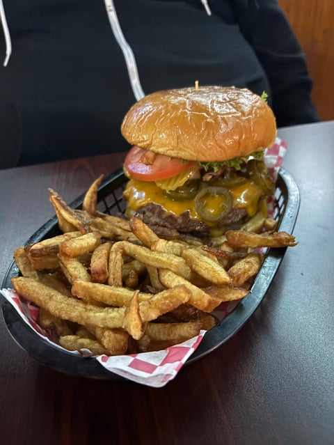 Gourmet cheeseburger with jalapeños and tomato, served with golden French fries on a dark plate with checkered paper