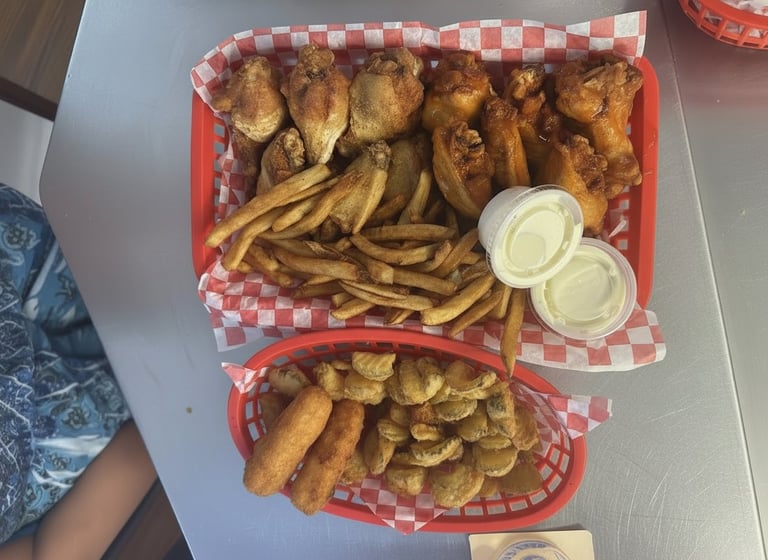 Red checkered takeout boxes filled with fried chicken pieces, french fries, and fried pickles with white sauce cups on white table