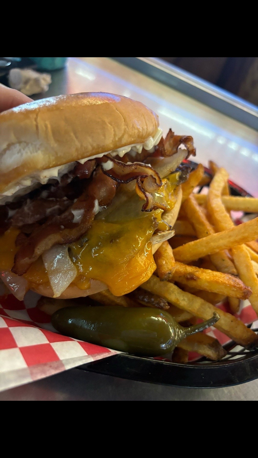 Cheeseburger with caramelized onions and melted cheddar cheese on a toasted bun, served with crispy fries and a jalapeño pepper on red checkered paper